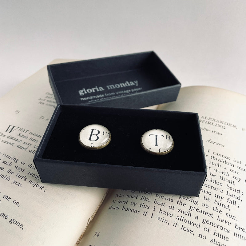 Cufflinks with 'B' and 'T' initials in a black box on an open book, branded 'Gloria Monday'.