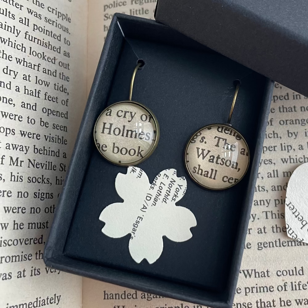 Pair of Holmes and Watson earrings with book page design in a black box on an open book