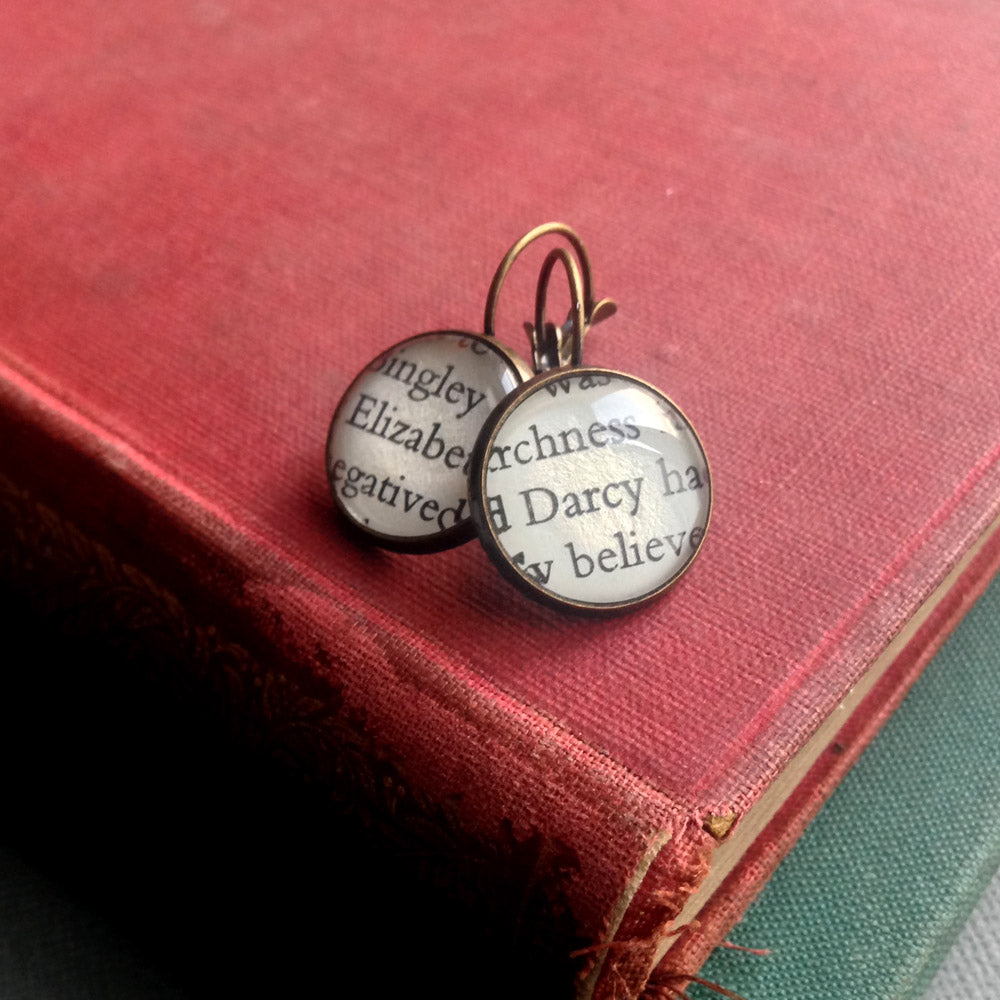 Bronze plated round earrings customised with glass and paper from pride and prejudice. one earring shows elizabeth and the other earring shows darcy. they are placed on a pile f antique books