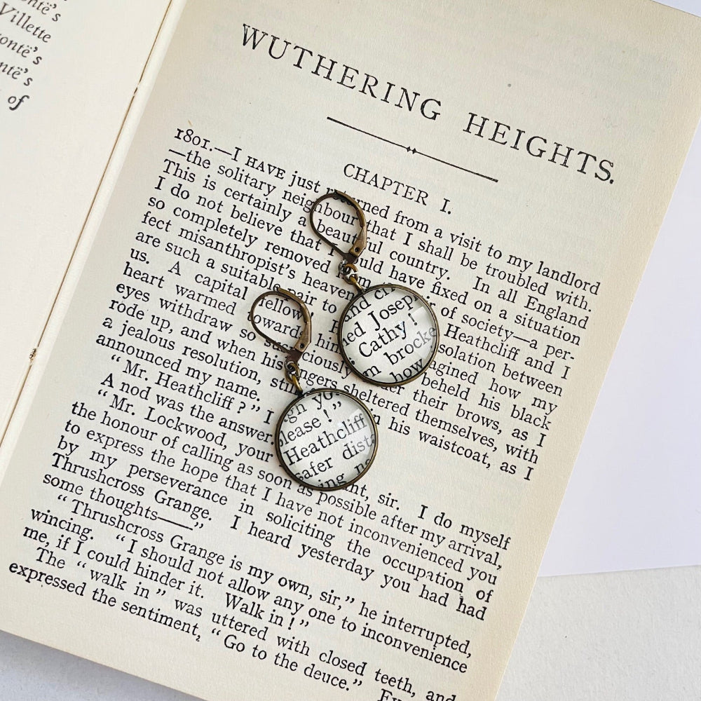 a pair of round bronze dangle earrings with 'Heathcliff' and 'Cathy' taken from real book pages. they are on an open copy of 'Wuthering Heights'