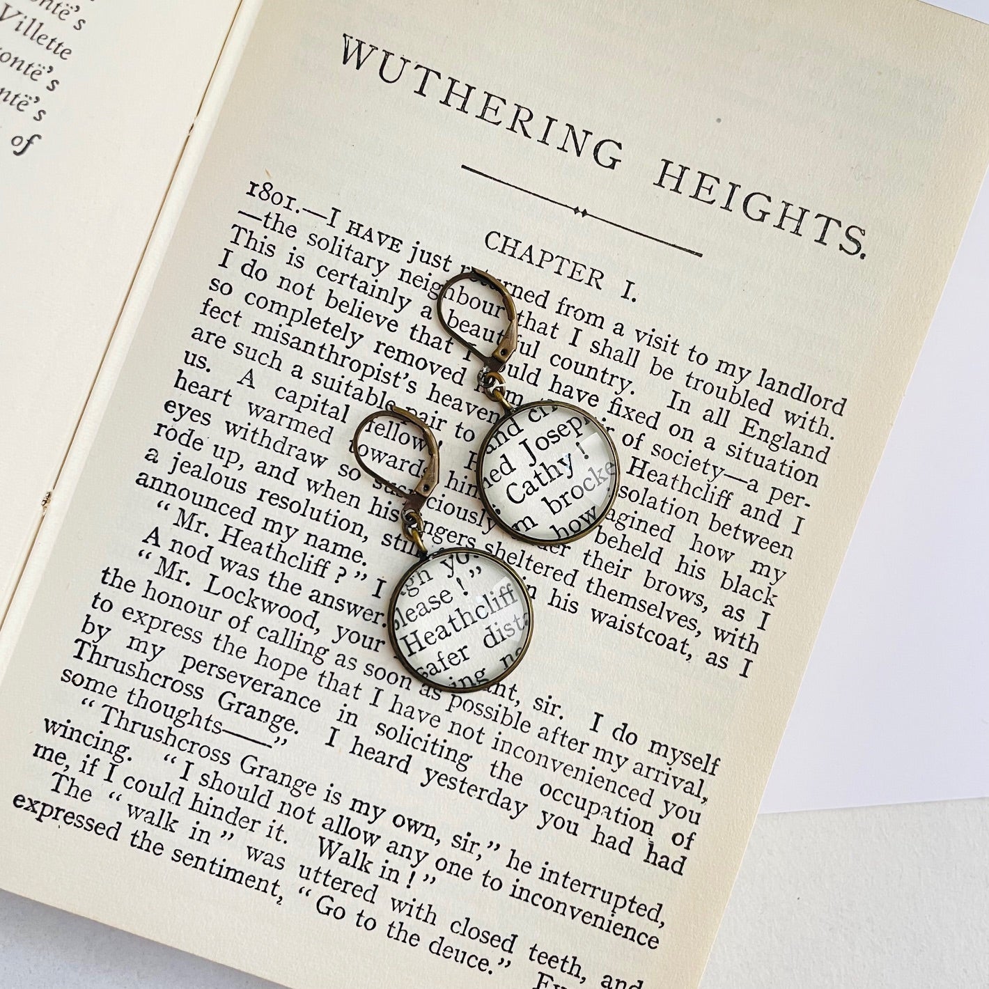 a pair of round bronze dangle earrings with 'Heathcliff' and 'Cathy' taken from real book pages. they are on an open copy of 'Wuthering Heights'
