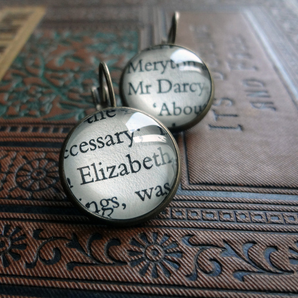 Bronze plated round earrings customised with glass and paper from pride and prejudice. one earring shows elizabeth and the other earring shows darcy. they are placed on an antique book that has an elaborate decorated cover