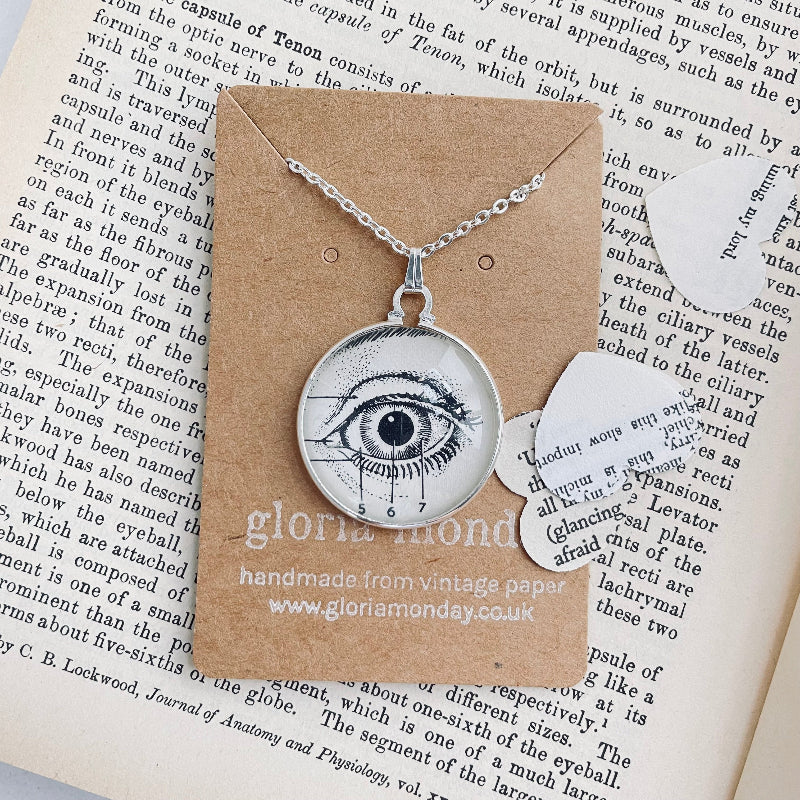 Eye-shaped pendant necklace on a card with 'gloria monday ' branding, placed on a book page.