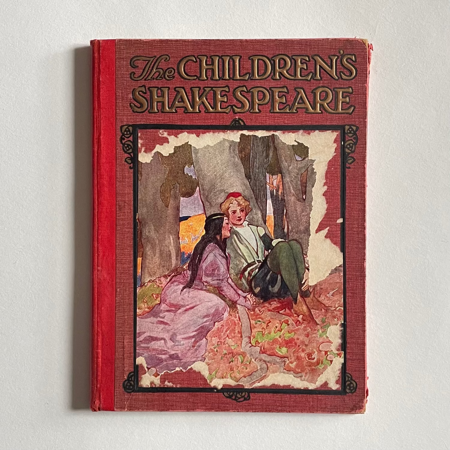 Book titled 'The Children's Shakespeare' with a red cover and illustration of two characters on a white background