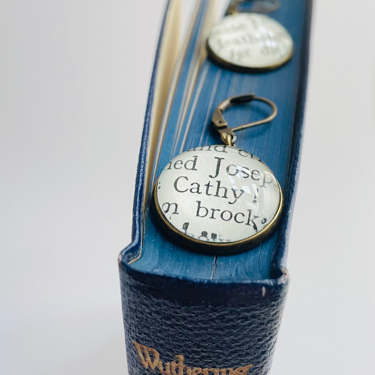 a pair of round bronze dangle earrings with 'Heathcliff' and 'Cathy' taken from real book pages. they are on an open copy of 'Wuthering Heights'